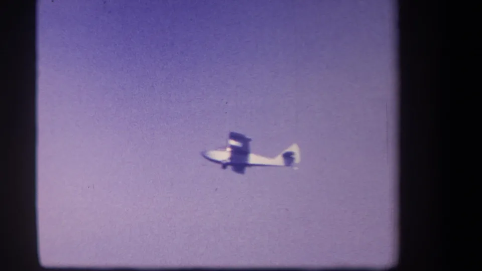 The white aircraft remains in flight near the lower center of the purple-tinted 8mm frame, with dust and soft focus visible.