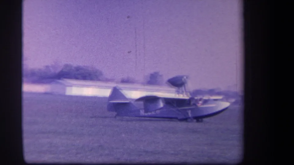 A small amphibious aircraft sits or taxis on grass, with low buildings in the background in 1959 Yucaipa footage.