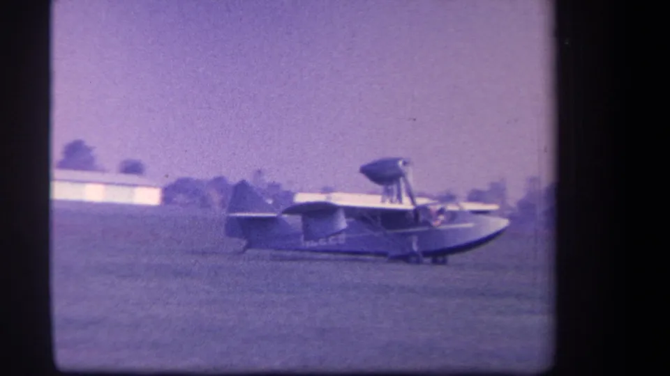 Side view of the distinctive amphibious plane on a grass field, with buildings and trees in the 1959 Yucaipa background.