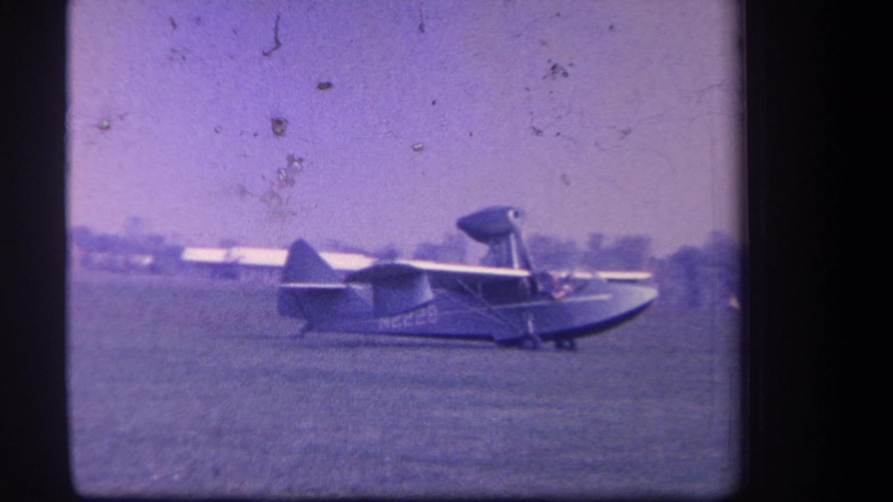 Small amphibian aircraft taxiing on a grass airfield with trees in the distance.