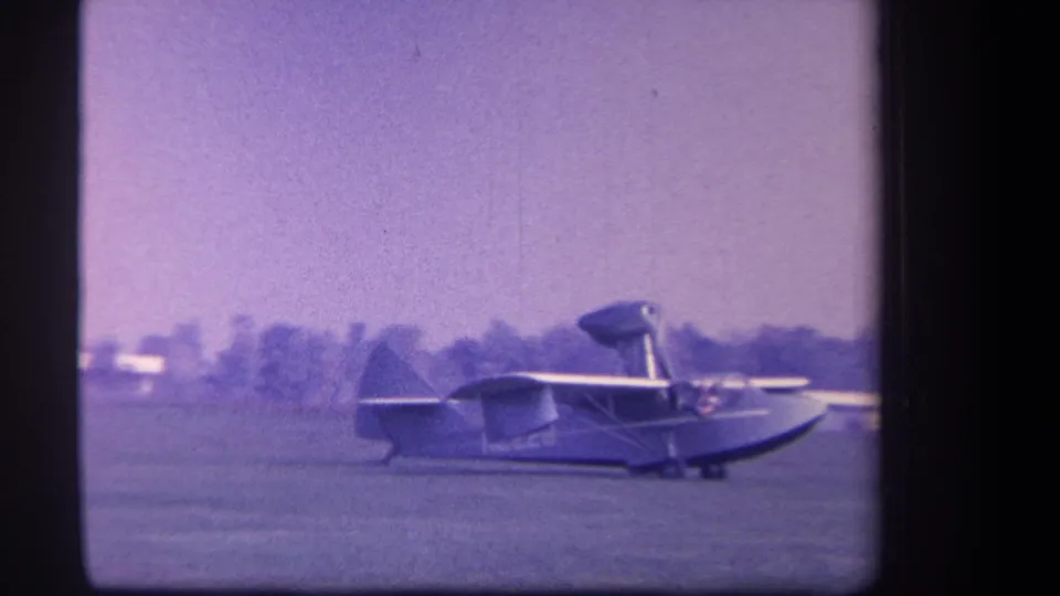 The Volmer amphibian aircraft appears farther across the Yucaipa grass airfield, with trees and a light sky behind it.
