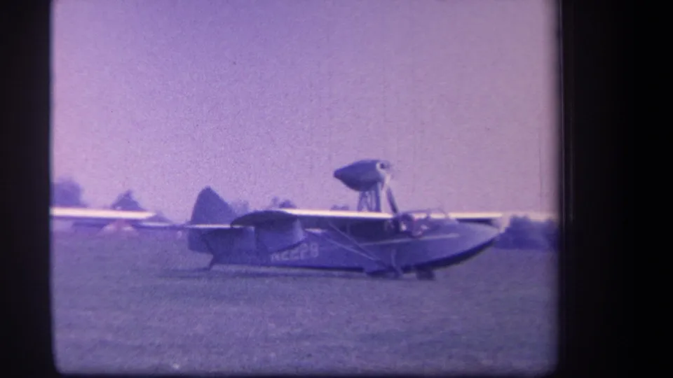 The Volmer amphibian aircraft taxis across the Yucaipa field in a soft 8mm frame with trees and open grass in the background.