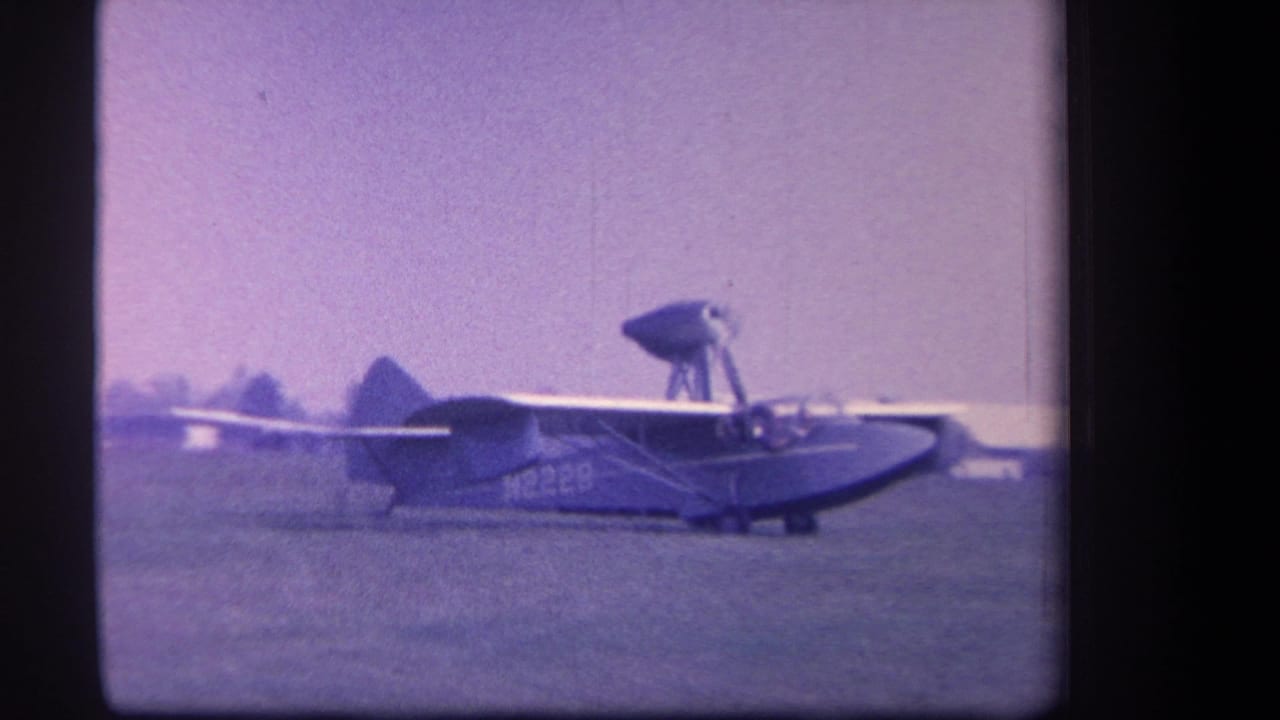 1959 Yucaipa home-movie frame showing a small amphibious aircraft moving across a grass airfield with distant trees behind it.