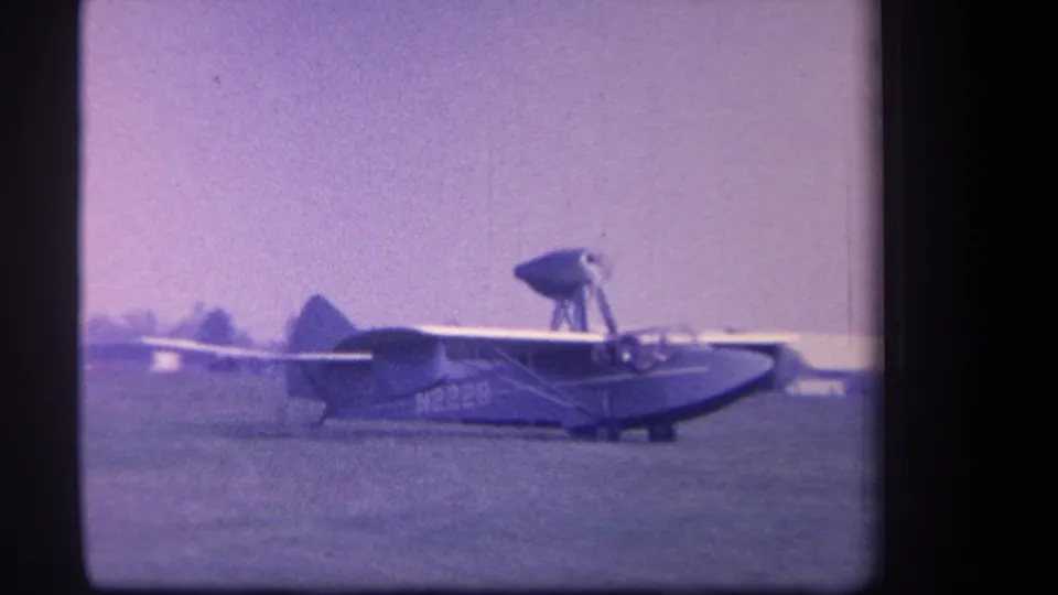 1959 Yucaipa home-movie frame showing a small amphibious aircraft moving across a grass airfield with distant trees behind it.