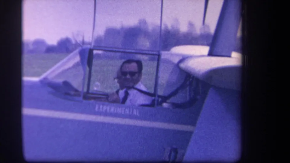 1959 home-movie frame showing a pilot in sunglasses and shirt with tie seated inside the small aircraft cockpit.