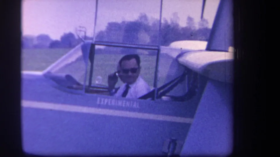 1959 Yucaipa home-movie frame showing a man in sunglasses seated in an experimental glider cockpit on a grassy field.