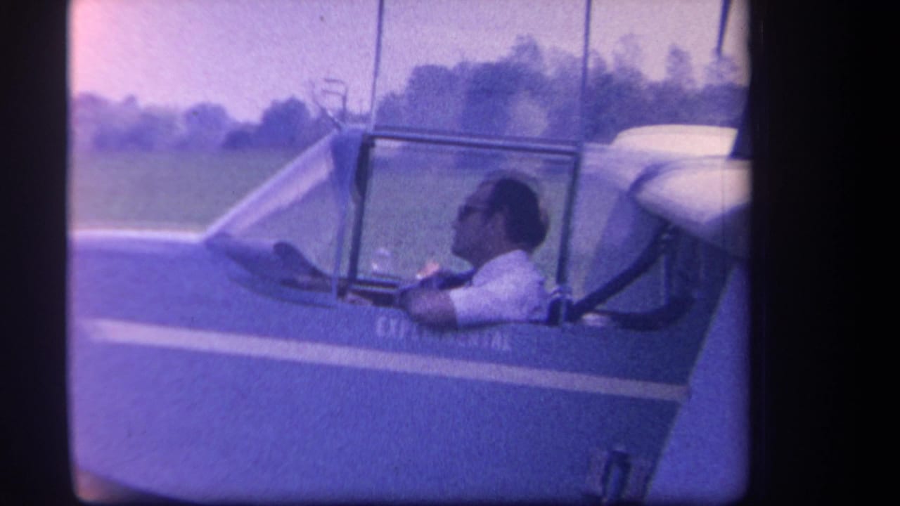 Man in sunglasses seated in a small aircraft cockpit on a grassy field.