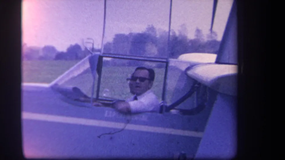 1959 Yucaipa-area frame of the pilot looking toward the camera from an experimental aircraft cockpit on a grassy airfield.