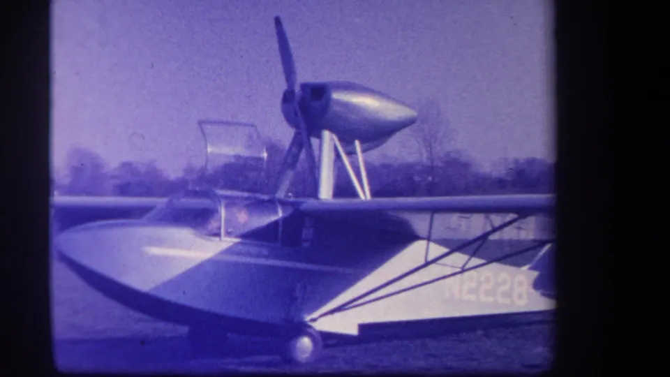 The 1959 8mm frame shows the amphibious aircraft from the side, with the nose, propeller, cockpit, wing struts, and registration area visible.