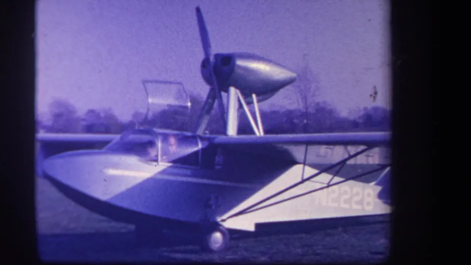 A purple-tinted 8mm frame shows the small amphibious aircraft parked in a field, with propeller, cockpit, landing gear, and partial registration visible.