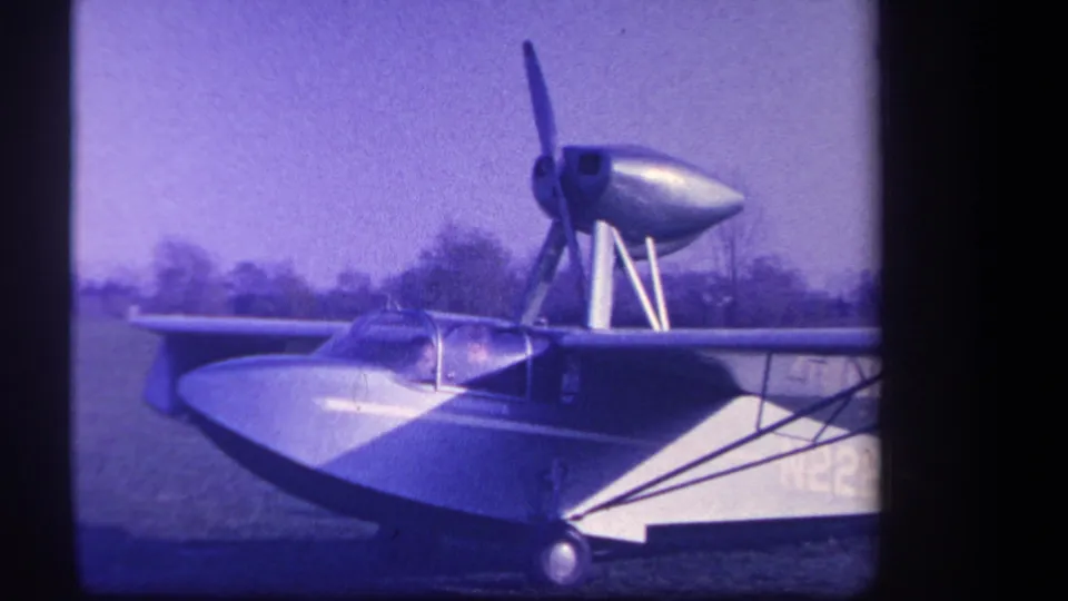 1959 Yucaipa 8mm frame with the aircraft side-on, propeller motion blur, cockpit canopy, wing struts, and distant trees.