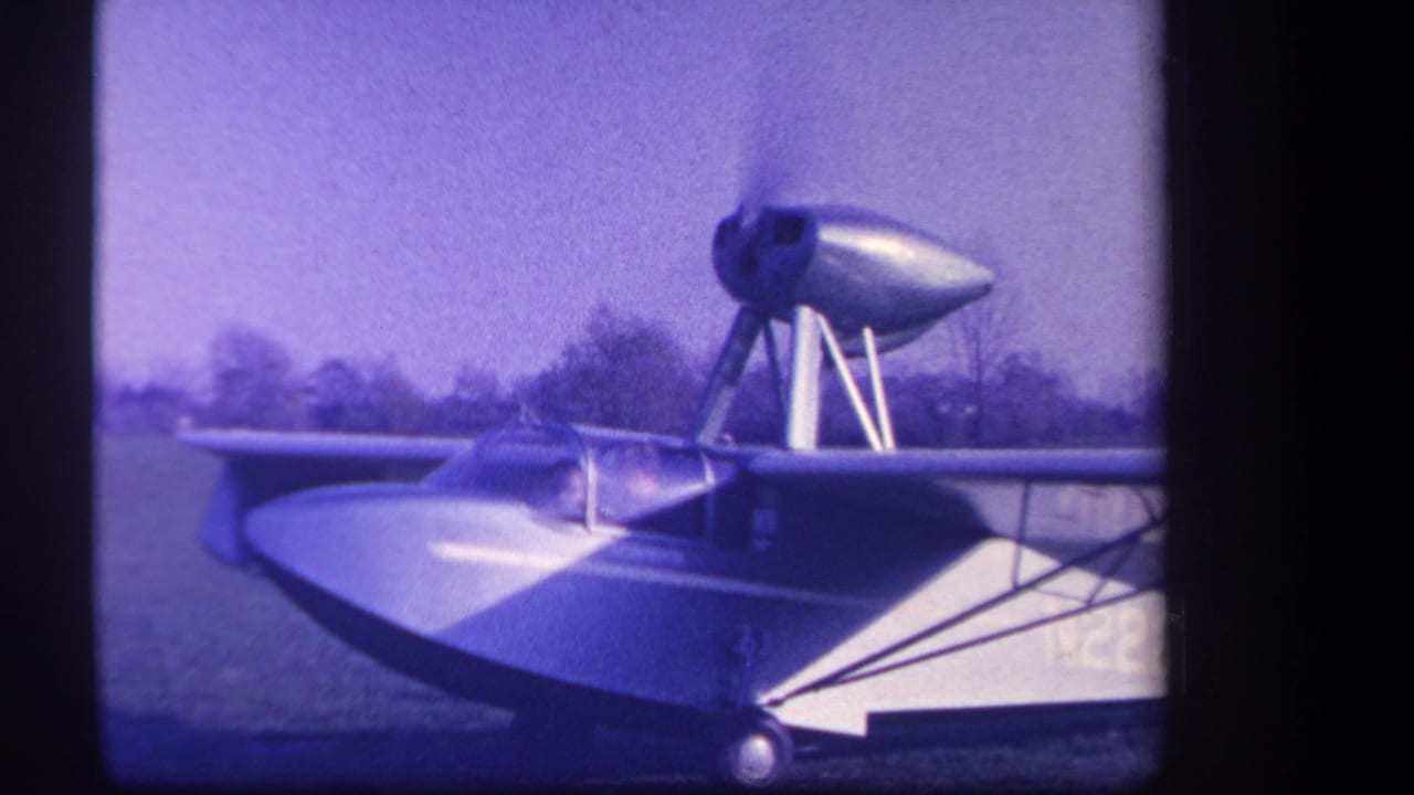 1959 Yucaipa-area 8mm frame showing an amphibious aircraft on grass with a high engine nacelle, wing struts, and purple film cast.