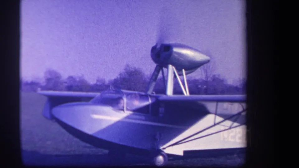 1959 Yucaipa-area 8mm frame showing an amphibious aircraft on grass with a high engine nacelle, wing struts, and purple film cast.