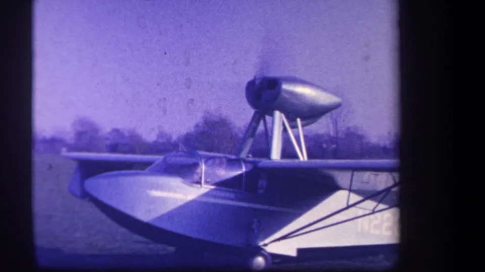 1959 8mm home-movie frame of the amphibious aircraft remaining stationary on grass, with cockpit glazing and a partial marking visible.