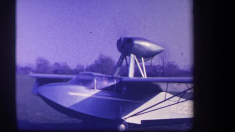 Final 1959 8mm frame of the amphibious aircraft in side view, showing the high-mounted engine, grass field, and partial aircraft marking.