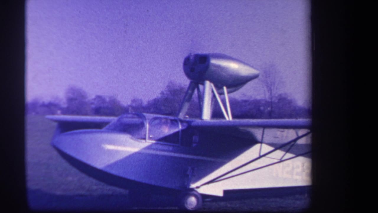 1959 Yucaipa-area 8mm frame showing a sleek amphibious aircraft with canopy, wheel, upper pod, and grass field behind it.
