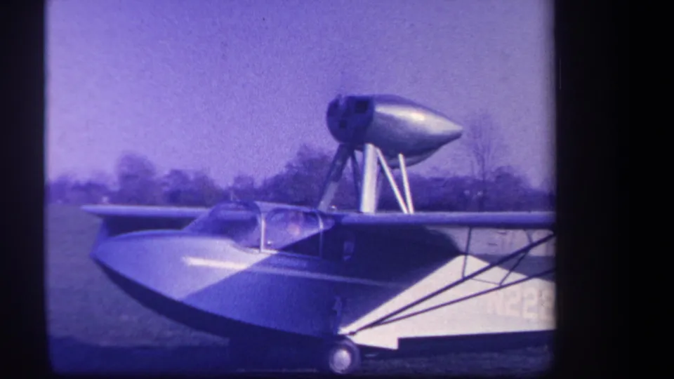 1959 Yucaipa-area 8mm frame showing a sleek amphibious aircraft with canopy, wheel, upper pod, and grass field behind it.