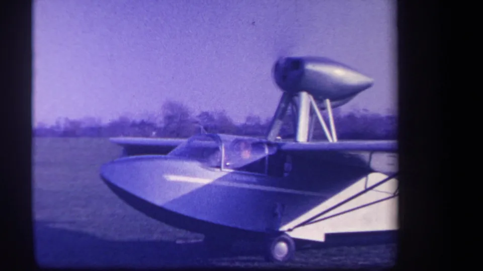 Vintage 8mm frame of the experimental amphibious aircraft on grass, with boat-like nose, canopy, struts, and upper pod visible.