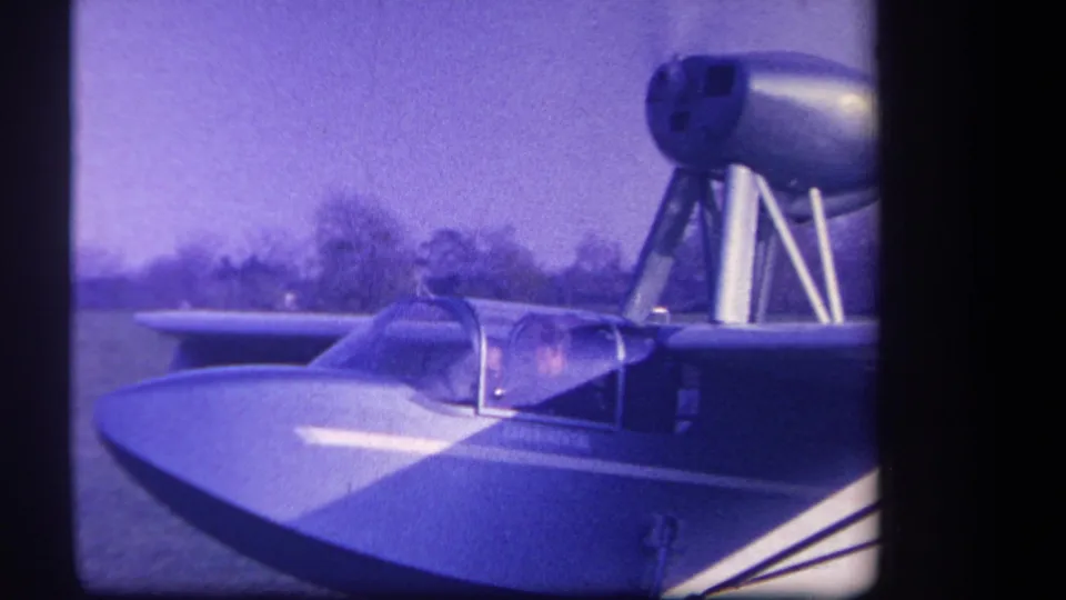 Close 1959 8mm view of the amphibious aircraft, emphasizing the rounded canopy, nose, and strut-mounted pod above the cockpit.