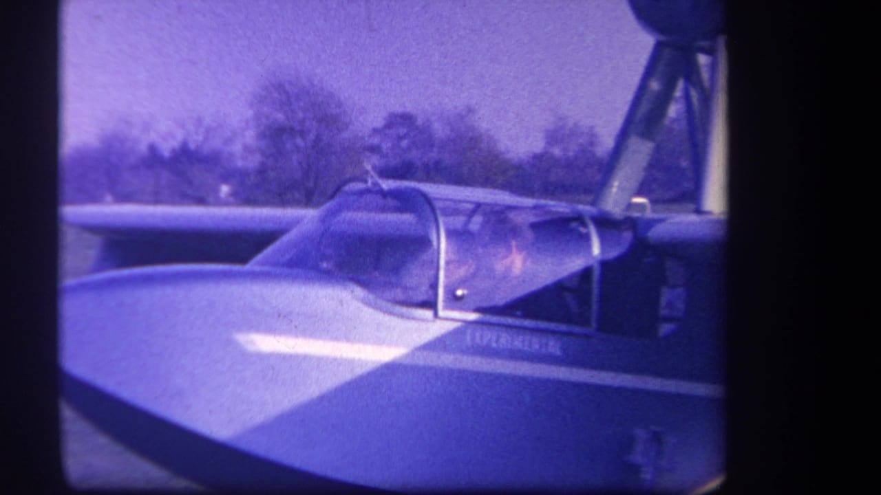 1959 8mm frame from Volmer Yucaipa, California, showing two people in the cockpit of a small experimental aircraft on a grassy field.
