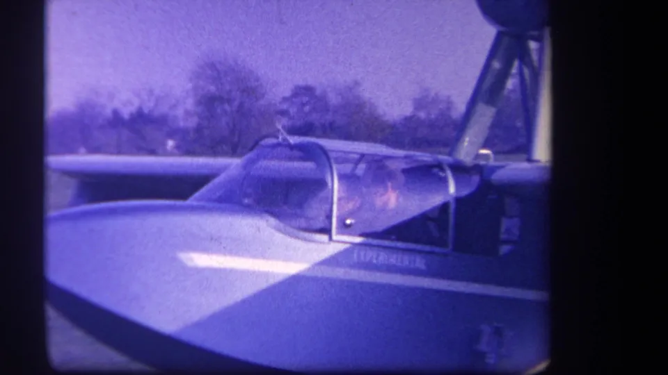 1959 8mm frame from Volmer Yucaipa, California, showing two people in the cockpit of a small experimental aircraft on a grassy field.