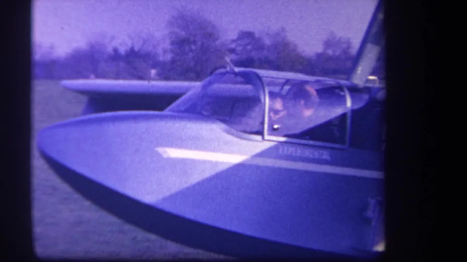 Blue-tinted 8mm frame from 1959 showing the small experimental aircraft at a grassy airfield, with the pilot visible in the cockpit.