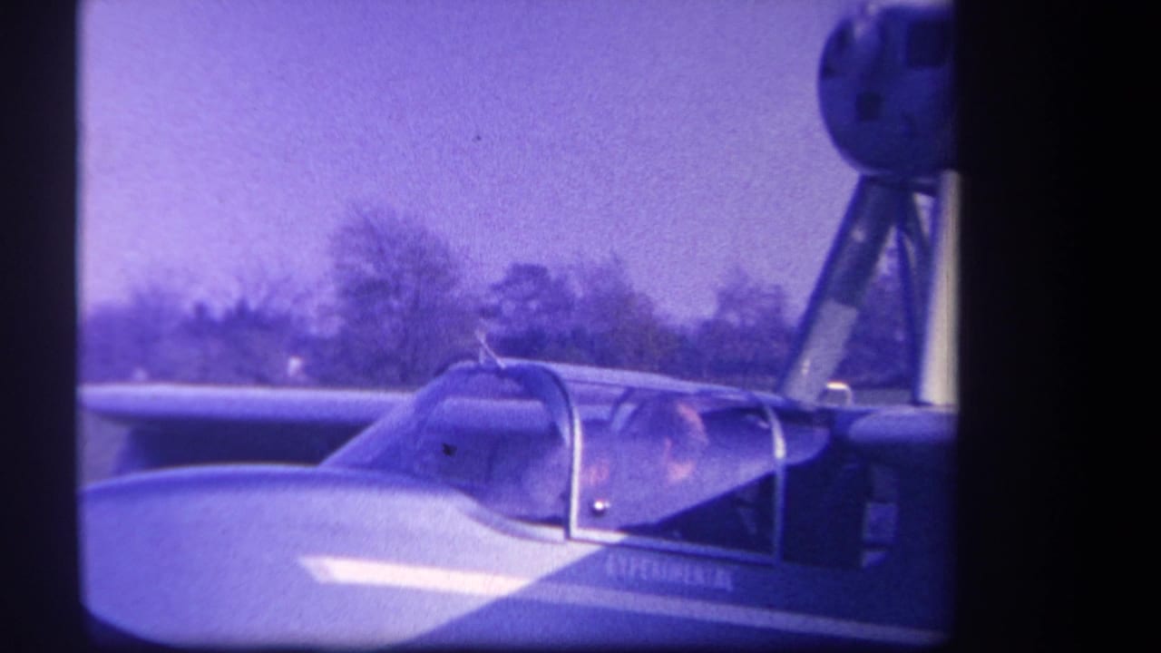 Small aircraft cockpit with a seated person, wing, tail structure, grass, and trees in view.