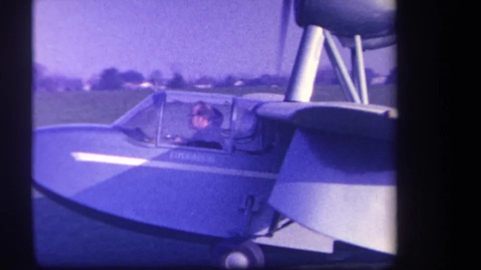 1959 Volmer Yucaipa frame with the pilot visible in the cockpit of a blue and white light aircraft on a grassy field.
