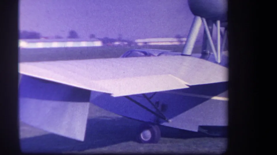 1959 Volmer Yucaipa frame showing the aircraft wing, support struts, landing wheel, and low buildings beyond the grass airfield.