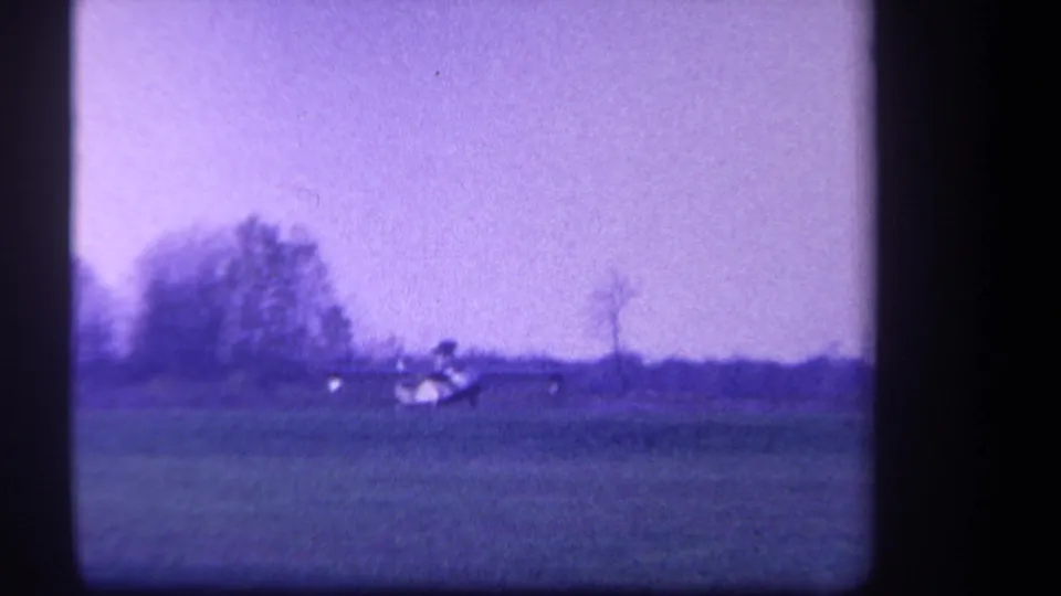 Final distant view of the small aircraft across the grassy field, with trees and sky dominating the frame.
