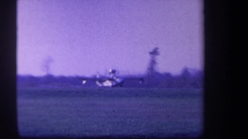Soft 8mm frame of a small plane across a rural field, with background trees and a strong purple tint.