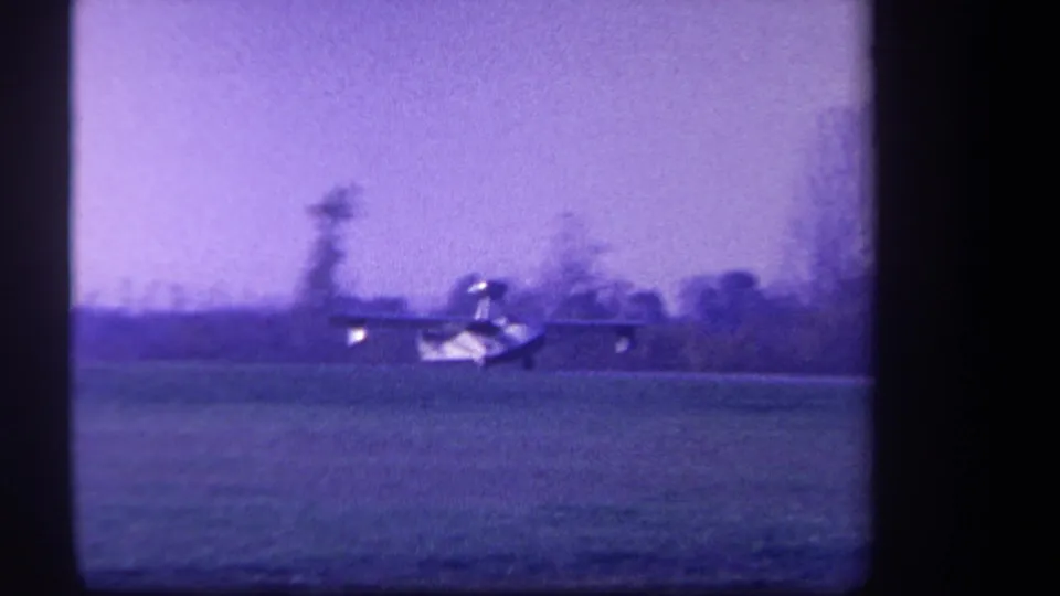The small aircraft is still visible on the grassy field, captured in grainy 8mm with a dark frame edge.