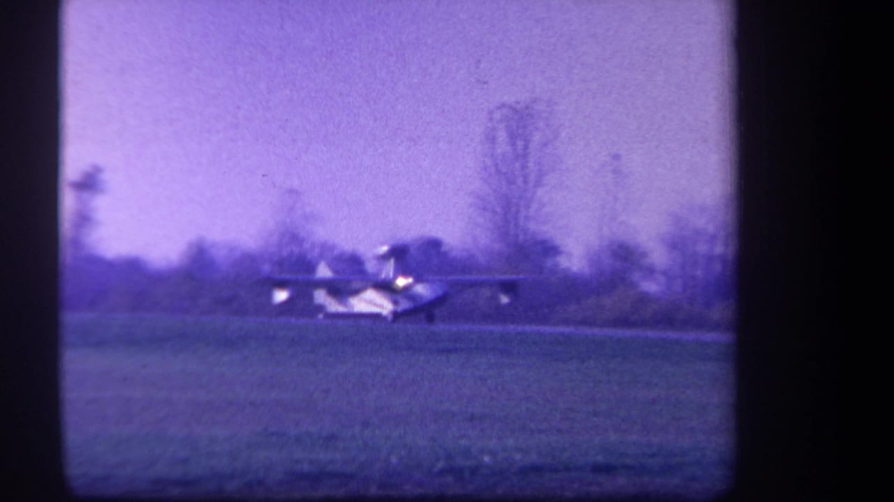 1959 home-movie frame from Yucaipa, California, showing a small pontoon-equipped airplane moving low across a grassy field.