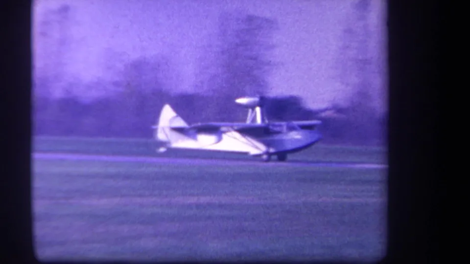 Close side view of the pontoon-equipped light aircraft passing across the grassy field in purple-tinted 8mm footage.