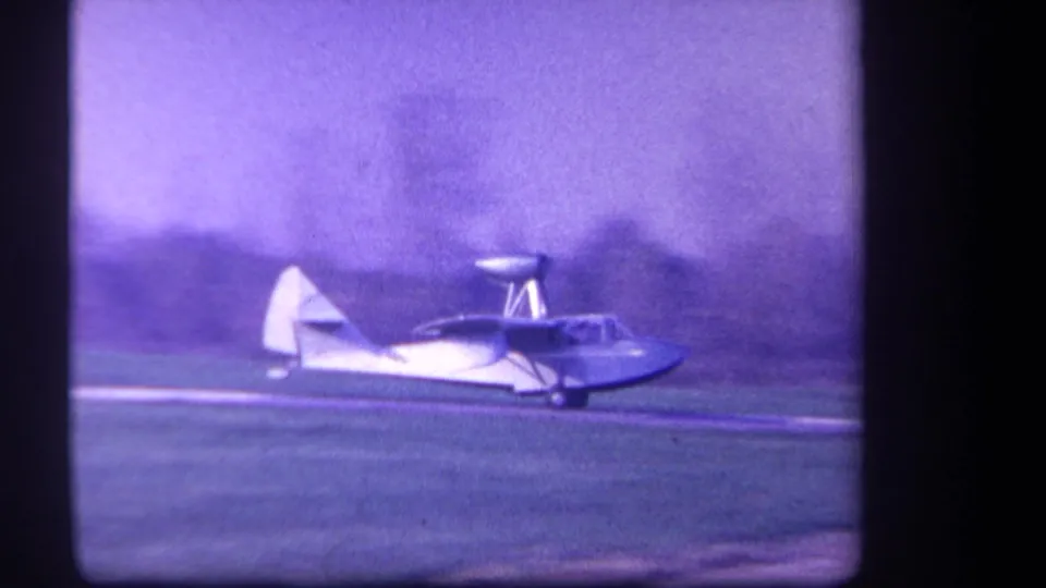 1959 Yucaipa-area home-movie frame showing a small white and red biplane rolling across a grass airfield under a hazy sky.