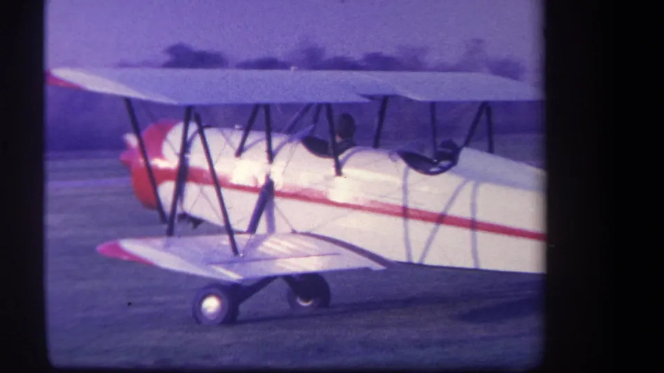 Close 8mm view of the white and red biplane as it passes near the camera on the Yucaipa-area grass airfield.