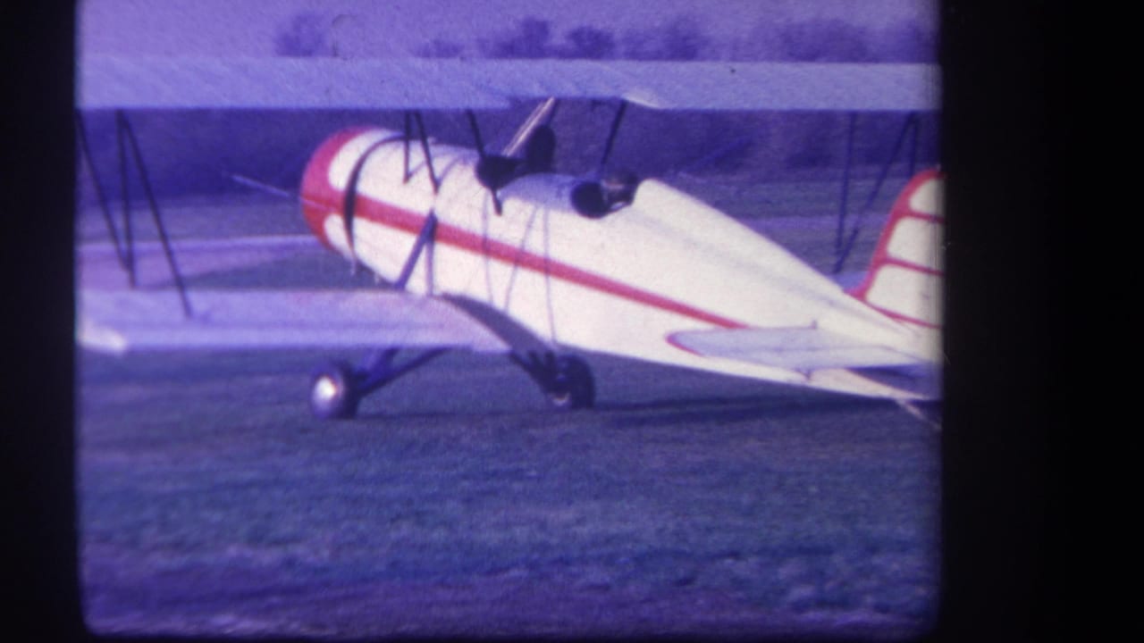 1959 Yucaipa home-movie frame showing a white red-striped biplane on a grass airfield with distant trees.