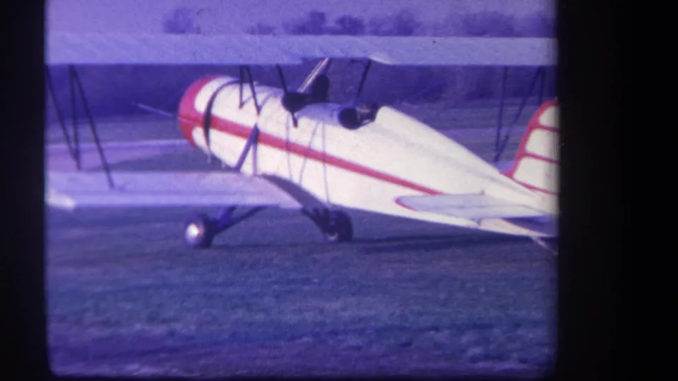 1959 Yucaipa home-movie frame showing a white red-striped biplane on a grass airfield with distant trees.