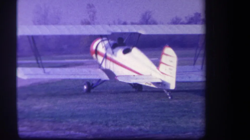 The red-striped biplane remains parked on the Yucaipa grass field, seen from a slightly rearward side angle.