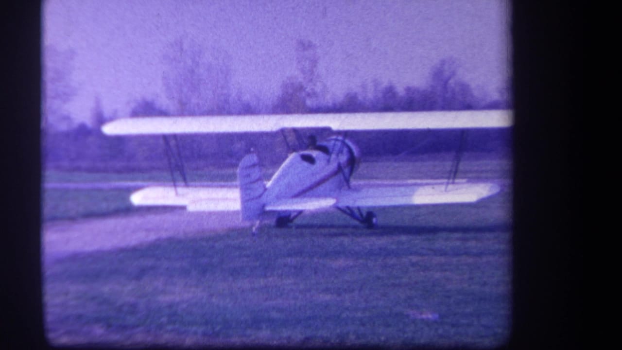 Red-and-white biplane on a grassy airfield seen through dark 8mm frame borders.