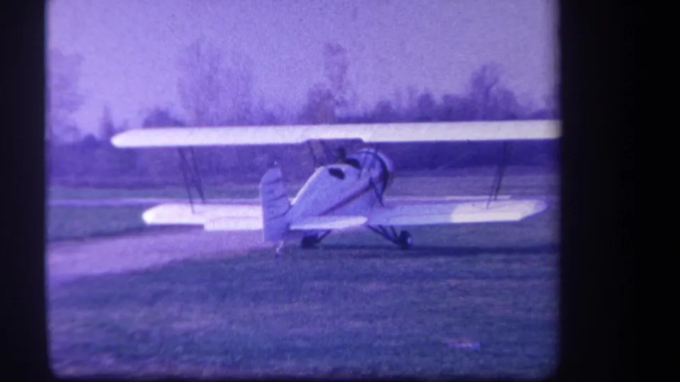1959 Yucaipa home-movie frame showing a red-and-white biplane on grass with a hazy tree line behind it.