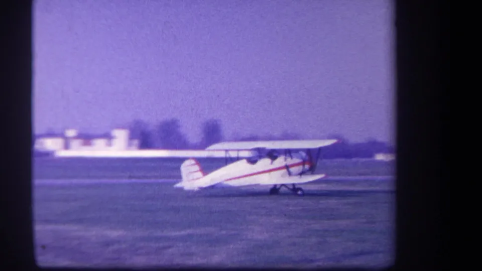 A 1959 8mm frame shows the biplane moving along the field with distant buildings and a hazy horizon behind it.