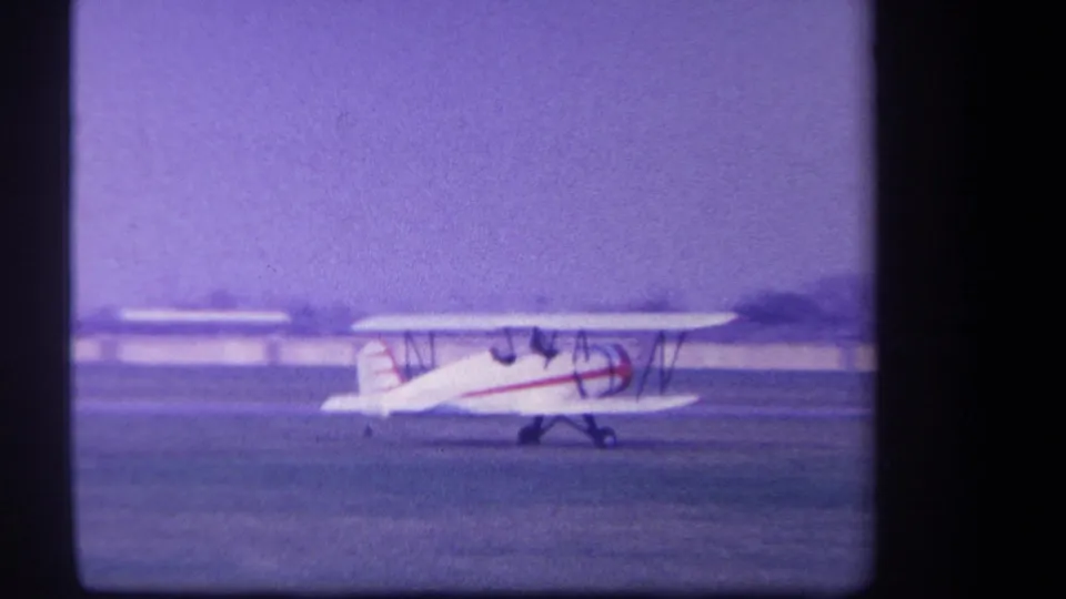 The red-and-white biplane continues its takeoff run across the Yucaipa field in soft, purple-shifted 8mm color.