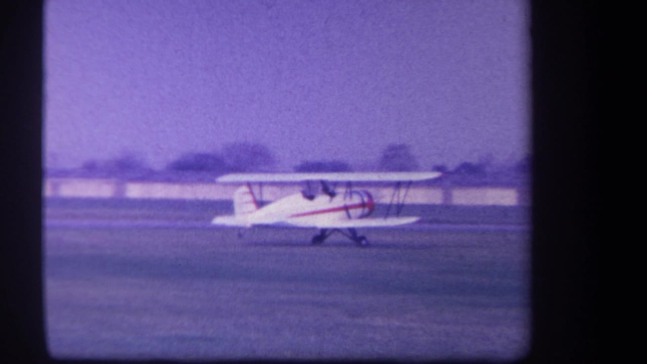 1959 Yucaipa-area 8mm frame showing a white biplane with red striping taxiing on grass under a pale purple sky.