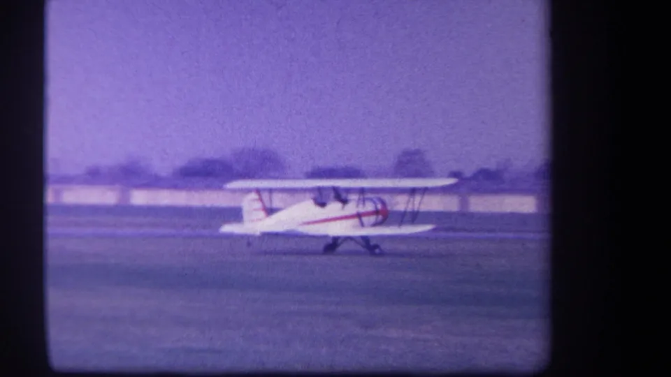 1959 Yucaipa-area 8mm frame showing a white biplane with red striping taxiing on grass under a pale purple sky.