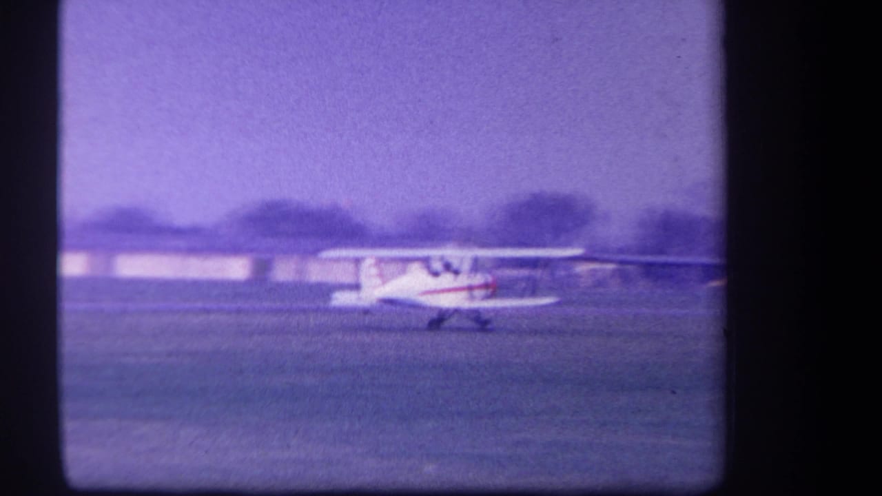 1959 Yucaipa-area 8mm frame with the white and red biplane taxiing across a grass airfield, low structures behind it.
