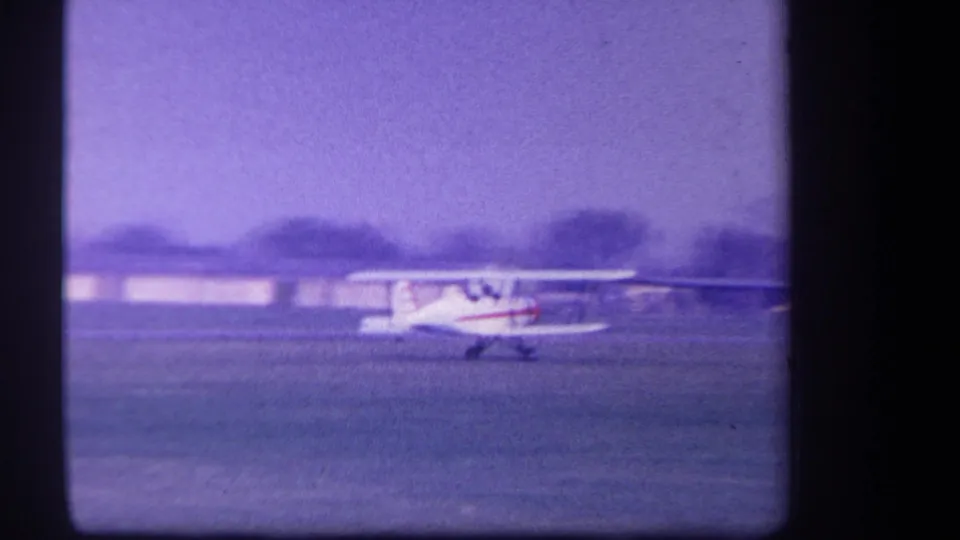 1959 Yucaipa-area 8mm frame with the white and red biplane taxiing across a grass airfield, low structures behind it.
