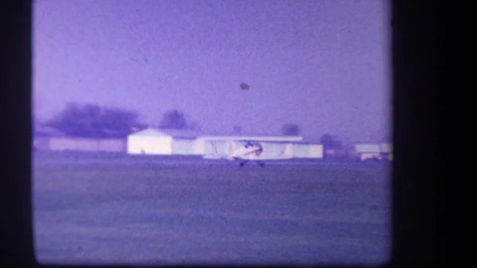 1959 Yucaipa-area 8mm frame showing the white and red biplane near light-colored airfield buildings on grass.
