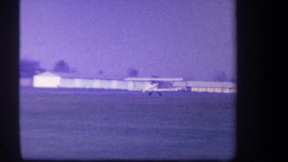 1959 Volmer Yucaipa 8mm frame showing a small biplane on or near a grass runway, with low airfield buildings and trees in the distance.