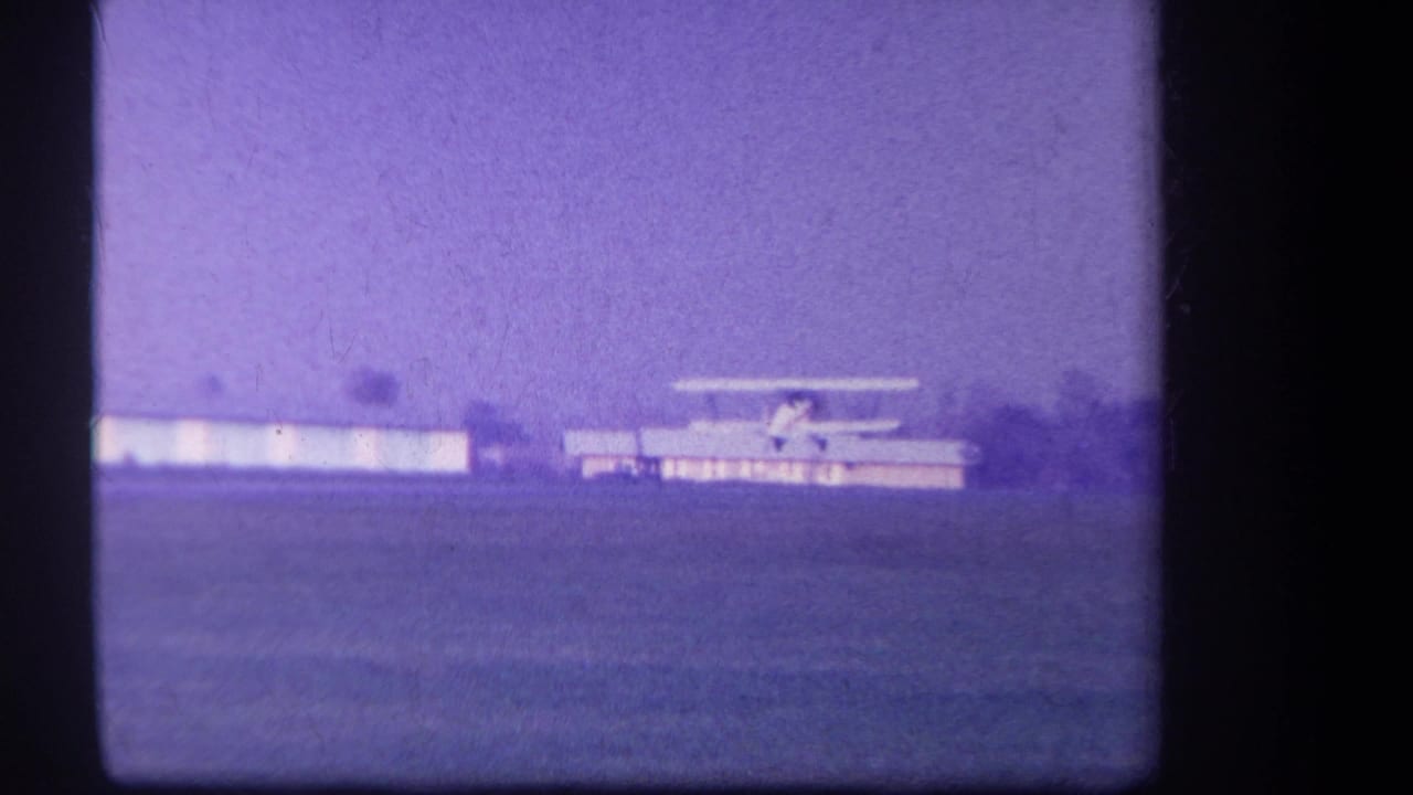 1959 Volmer Yucaipa frame with a small biplane gaining speed across a rural airfield, backed by low buildings and a distant tree line.